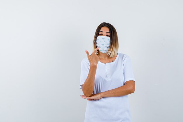 Young girl pointing at herself with index finger, holding hand under elbow in white t-shirt , mask and looking serious , front view.