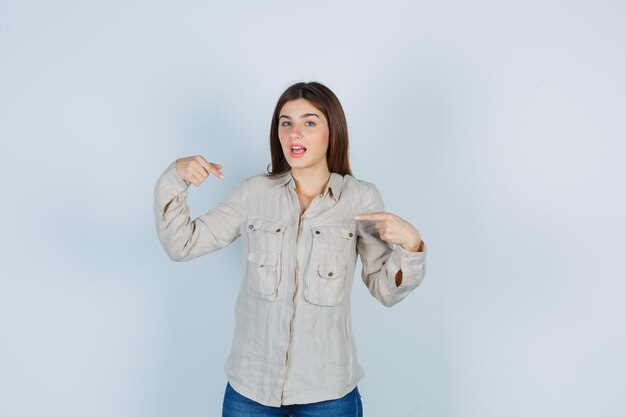 Young girl pointing at herself with index fingers in beige shirt, jeans and looking excited , front view.