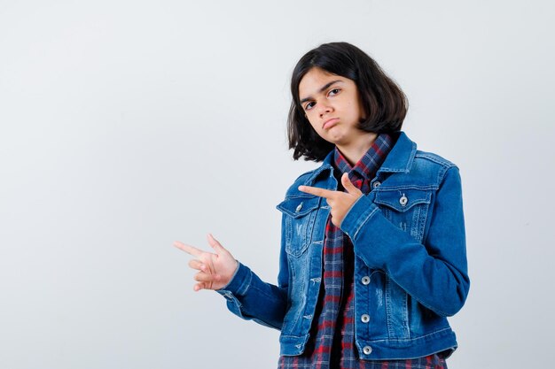체크 셔츠와 진 재킷을 입고 왼쪽을 가리키며 진지한 표정을 짓고 있는 어린 소녀