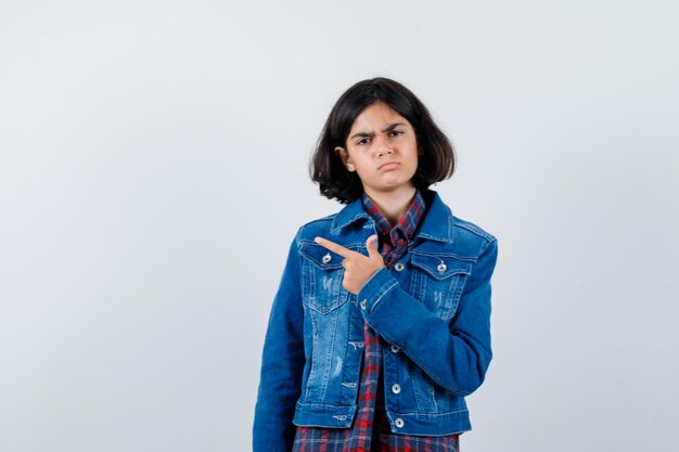 Молодая девушка указывает влево указательным пальцем в клетчатой рубашке и джинсовой куртке и серьезно выглядит, вид спереди.