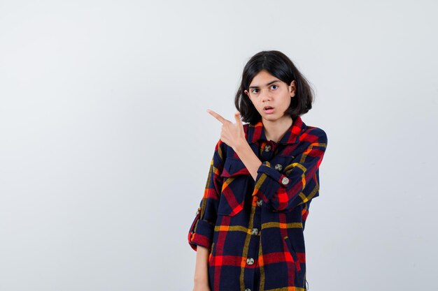 Young girl pointing left with index finger in checked shirt and looking surprised. front view.