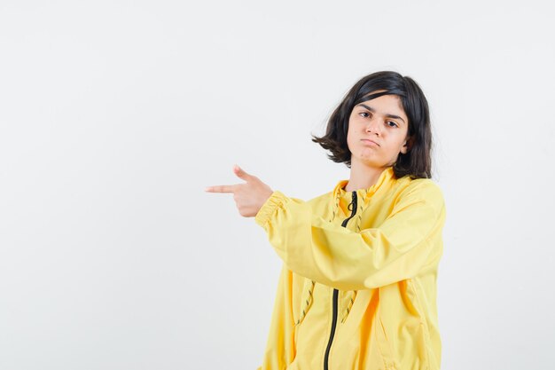 Молодая девушка в желтой куртке-бомбардировке показывает пальцем влево и серьезно выглядит