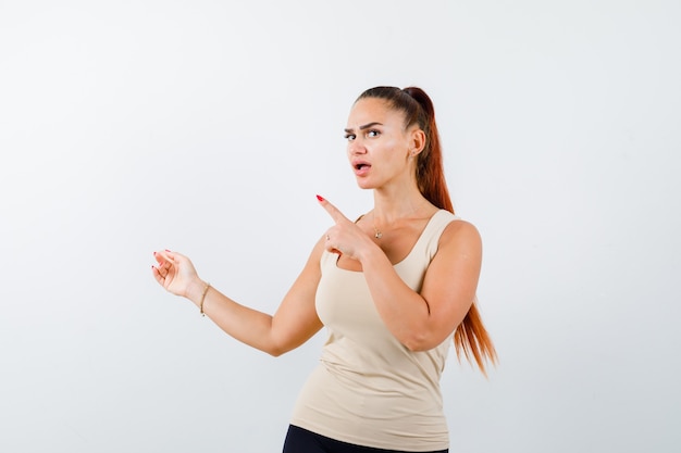 Young girl pointing left with index fingers in beige top, black pants and looking surprised