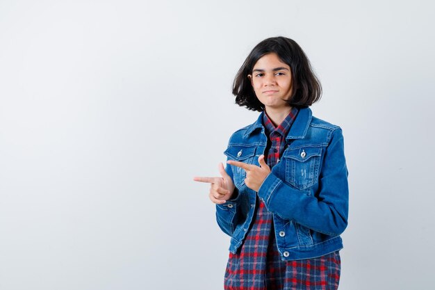 Молодая девушка указывает влево указательными пальцами в клетчатой рубашке и джинсовой куртке и выглядит мило, вид спереди.
