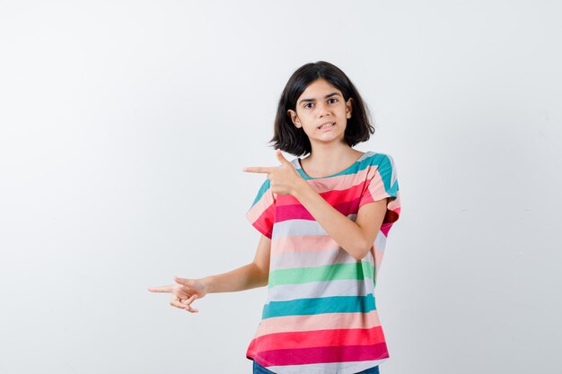 Молодая девушка указывает влево указательными пальцами в красочной полосатой футболке и выглядит серьезной, вид спереди.