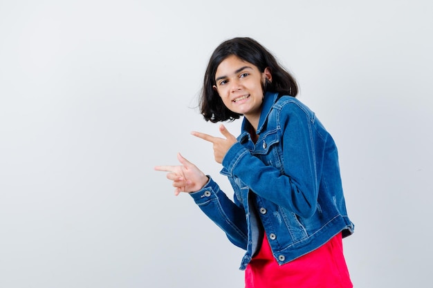 Young girl pointing left with index fingers in red t-shirt and jean jacket and looking happy. front view.