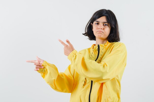 Молодая девушка в желтой куртке-бомбардировке, указывающая влево указательными пальцами, выглядит серьезной.
