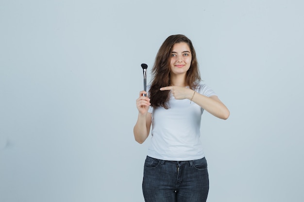 Young girl pointing makeup brush in t-shirt, jeans and looking confident , front view.