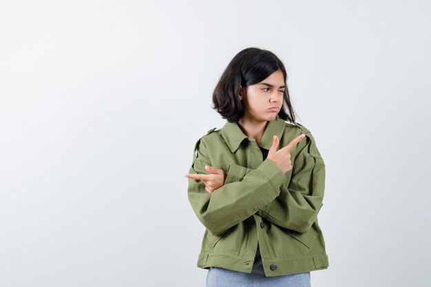 灰色のセーター、カーキ色のジャケット、ジーンズのパンツで反対方向を指している若い女の子と焦点を合わせて、正面図。