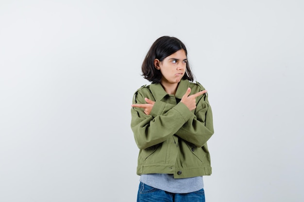 Giovane ragazza che punta in direzioni opposte in maglione grigio, giacca color kaki, pantaloni di jeans e sembra seria. vista frontale.