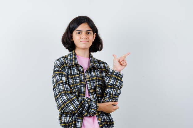 Young girl pointing right and holding one hand on elbow in checked shirt and pink t-shirt and looking cute. front view.