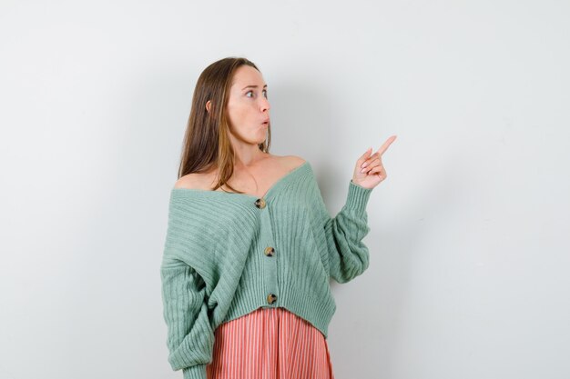 Young girl pointing right with index finger in knitwear, skirt and looking surprised , front view.