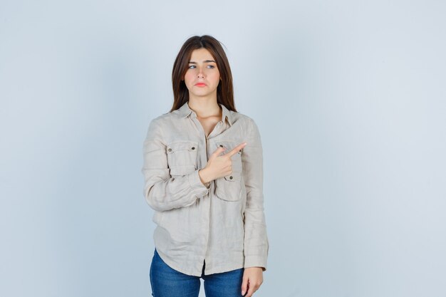 Young girl pointing right with index finger, looking away in beige shirt, jeans and looking sad. front view.