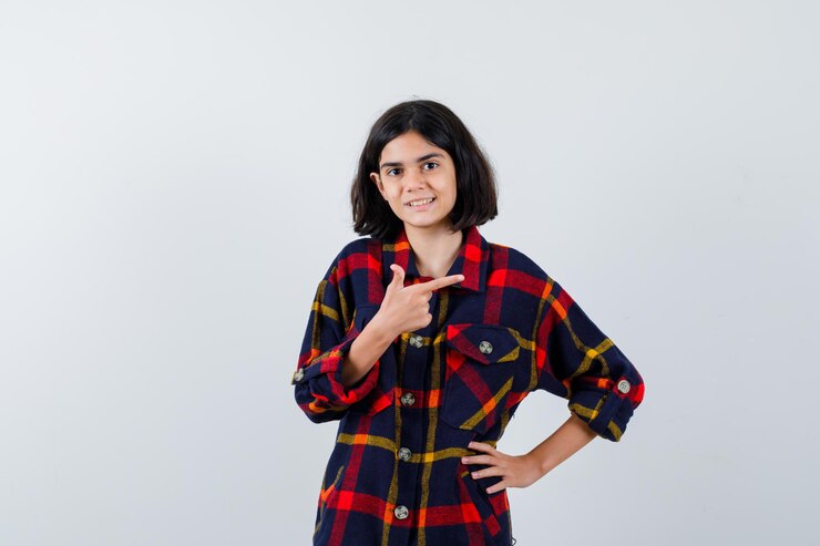 मुफ़्त फोटो young girl pointing right with index finger while holding hand on waist in checked shirt and looking happy , front view.