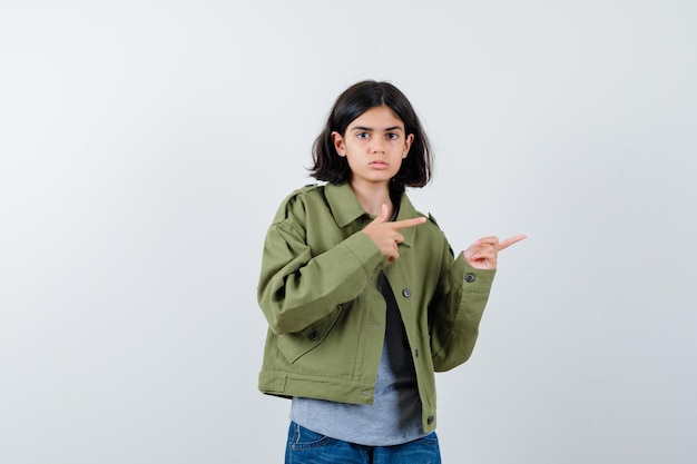 Giovane ragazza che punta a destra con l'indice in maglione grigio, giacca color kaki, pantaloni di jeans e sembra seria, vista frontale.