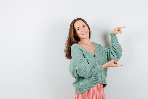 Young girl pointing right with index fingers in knitwear, skirt and looking happy. front view.