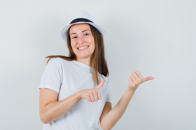 Young girl pointing to the side with thumbs in white t-shirt, hat and looking glad , front view.