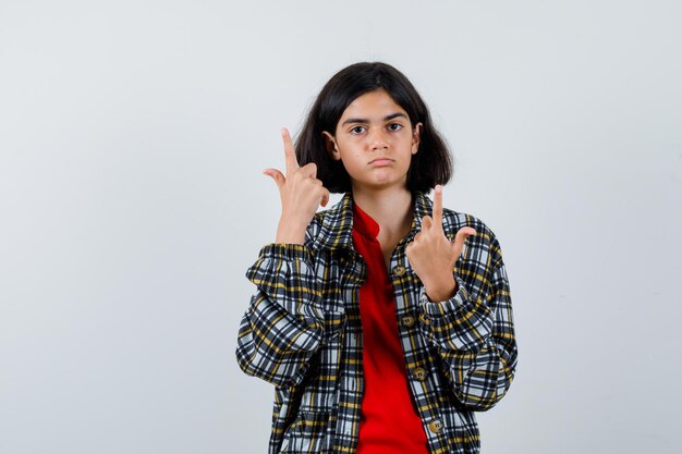 Young girl pointing up in checked shirt and red t-shirt and looking serious. front view.