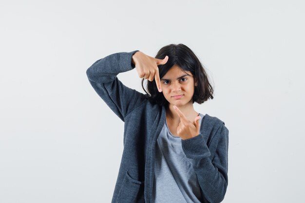 Young girl pointing up and down with index fingers in light gray t-shirt and dark grey zip-front hoodie and looking serious