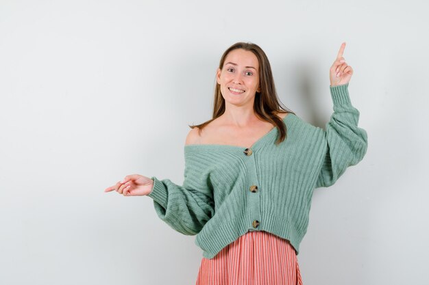 Young girl pointing up and left with index fingers in knitwear, skirt and looking happy. front view.