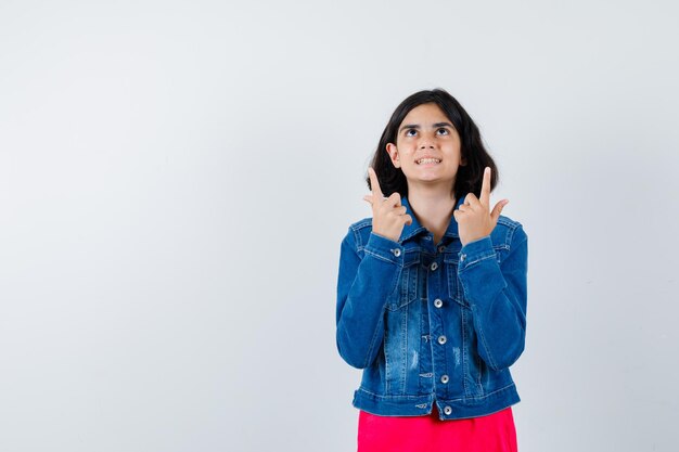 Young girl pointing up in red t-shirt and jean jacket and looking happy , front view.