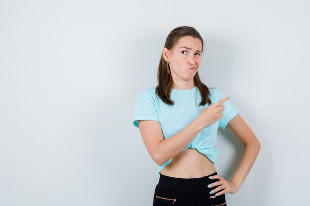 Young girl pointing up, with hand on waist, twisting mouth to the side in turquoise t-shirt, pants and looking dissatisfied , front view.