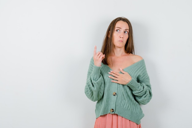 Young girl pointing up with index finger, holding hand over chest in knitwear, skirt and looking pensive. front view.