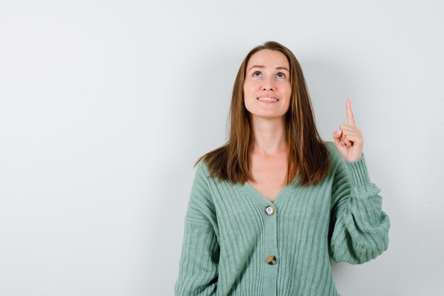 Young girl pointing up with index finger in knitwear, skirt and looking cheery. front view.