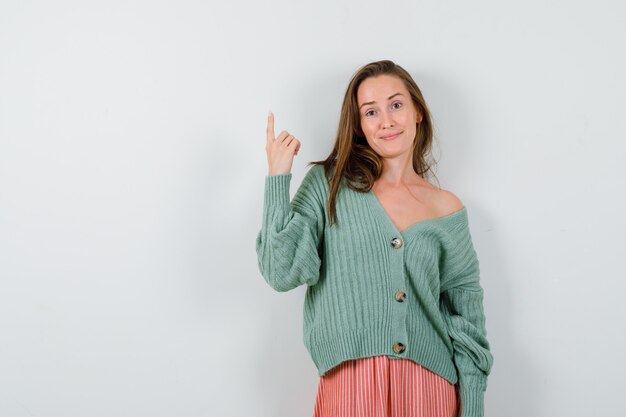 Young girl pointing up with index finger in knitwear, skirt and looking happy. front view.