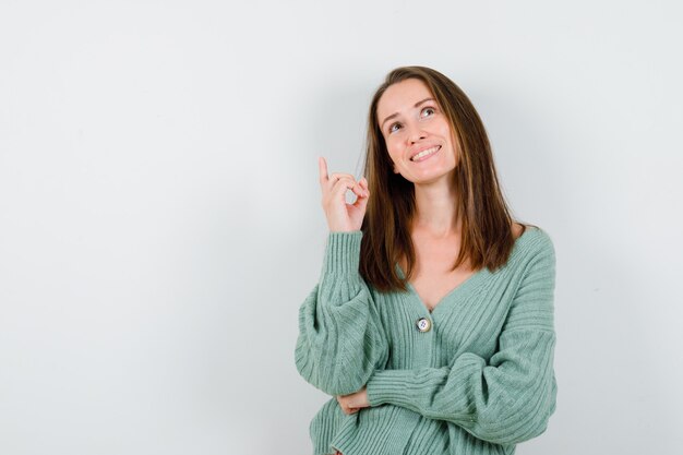 Young girl pointing up with index finger, looking upward in knitwear, skirt and looking happy. front view.