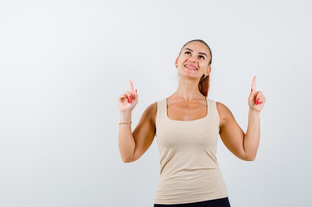 Young girl pointing up with index fingers in beige top, black pants and looking cheery , front view.