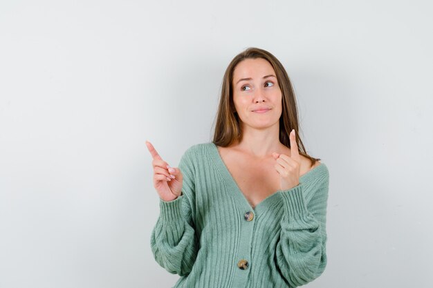 Young girl pointing up with index fingers, looking away in knitwear and looking happy. front view.