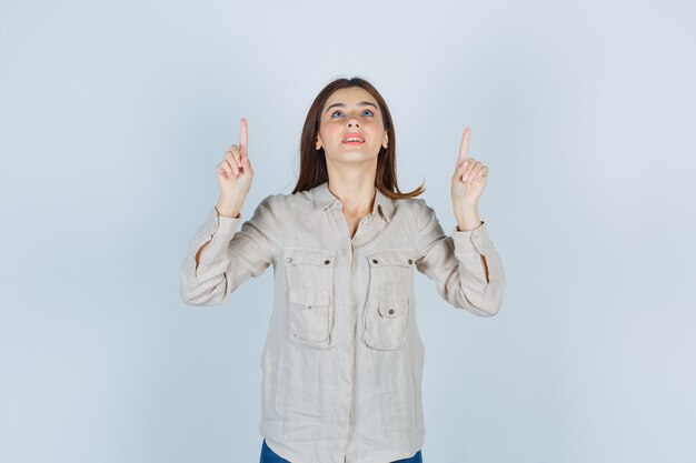 Young girl pointing up with index fingers, looking upward in beige shirt, jeans and looking cute , front view.