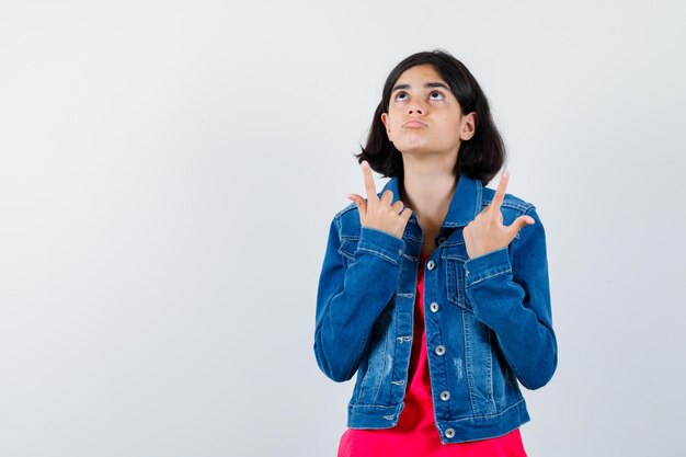 Young girl pointing up with index fingers in red t-shirt and jean jacket and looking cute. front view.