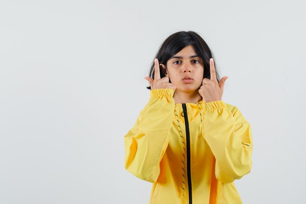Young girl pointing up with index fingers in yellow bomber jacket and looking serious