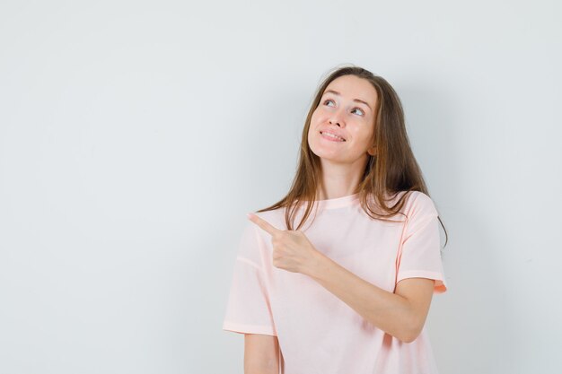 Молодая девушка указывая на верхний левый угол в розовой футболке и выглядит мило, вид спереди.