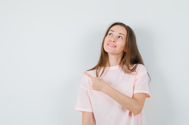 Young girl pointing at upper left corner in pink t-shirt and looking cute , front view.