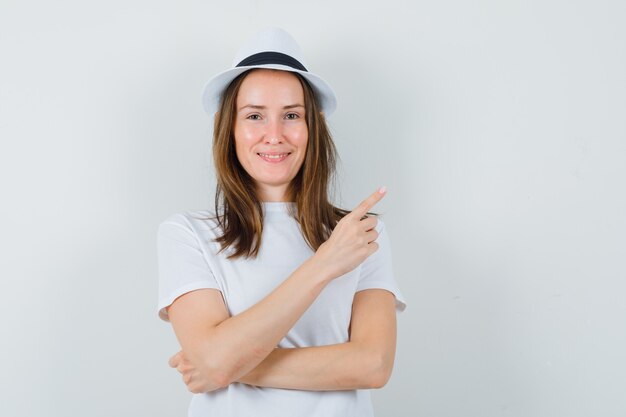 Молодая девушка указывая на верхний правый угол в белой футболке, шляпе и уверенно глядя, вид спереди.