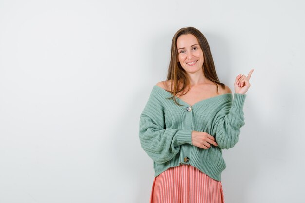 Young girl pointing upper right side with index finger in knitwear, skirt and looking happy. front view.