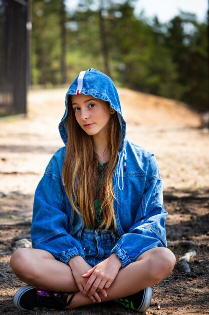 A young girl poses in a denim jacket with a hood against the of a forest sitting