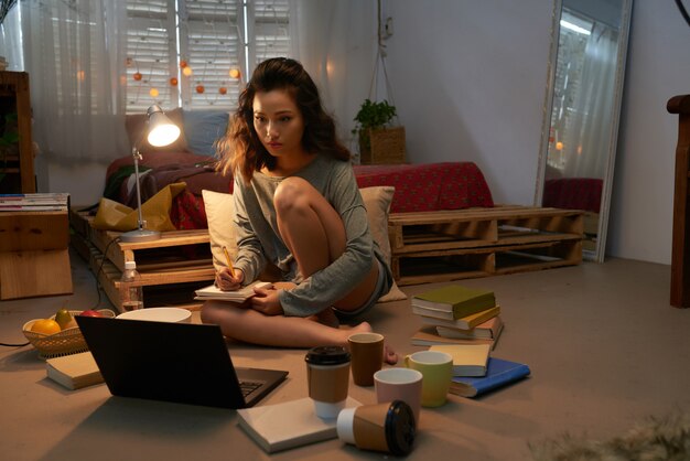 Young girl preparing for exam seated on the floor of her dorm room surrounded by laptop, books and empty cups