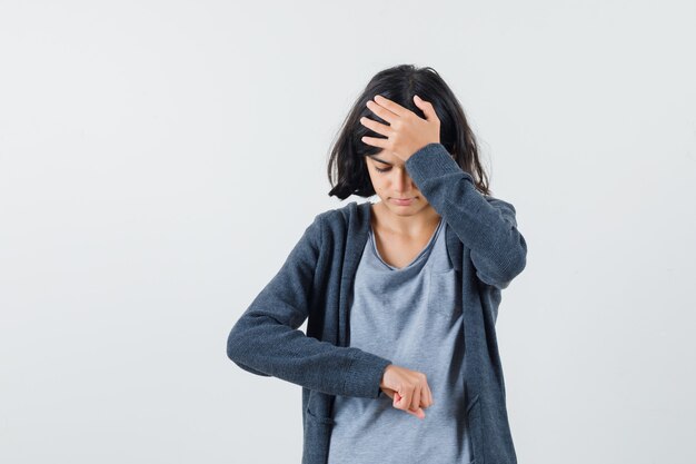 Young girl pretending like looking at clock while putting hand on forehead in light gray t-shirt and dark grey zip-front hoodie and looking stressed