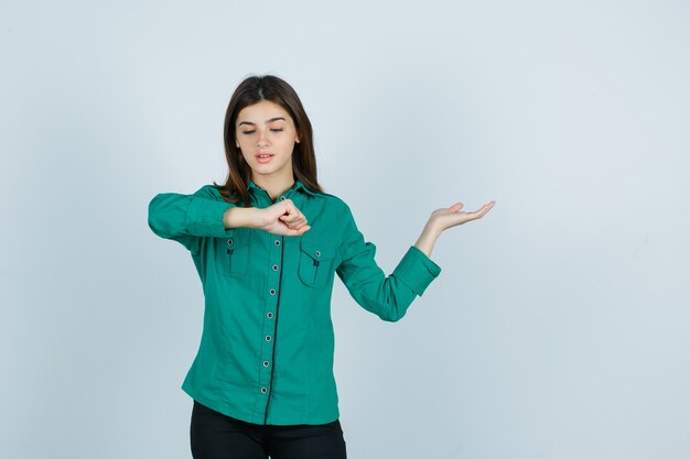 Молодая девушка делает вид, будто смотрит на часы на запястье, разводит ладонь в зеленой блузке, черных штанах и смотрит сосредоточенно, вид спереди.