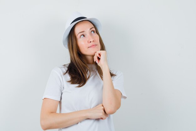 Young girl propping chin on fist in white t-shirt, hat and looking dreamy. front view.