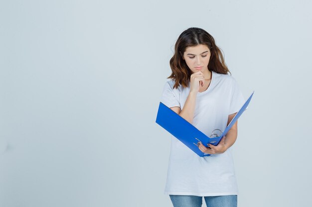 Young girl propping chin on hand, looking into folder in white t-shirt and looking pensive. front view.