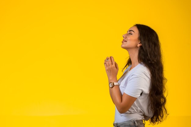 Young girl puts her hands under her chin and prays on yellow background. 