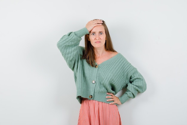 Young girl putting hand on forehead, holding hand on waist in knitwear, skirt and looking annoyed. front view.