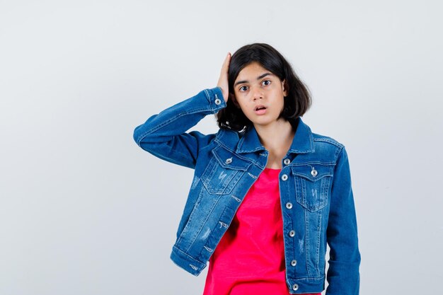 Young girl putting hand on head in red t-shirt and jean jacket and looking surprised. front view.