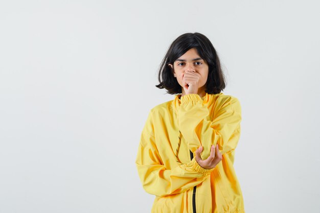 Young girl putting hand on mouth, clenching fist and thinking about something in yellow bomber jacket and looking pensive