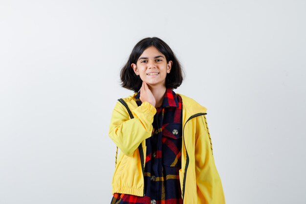 Young girl putting hand on neck in checked shirt and yellow jacket and looking happy. front view.